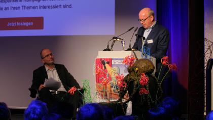 Peter Wedde am Redner.innenpult. Neben ihm sitzt Sönke Hilbrans auf einem Stuhl.